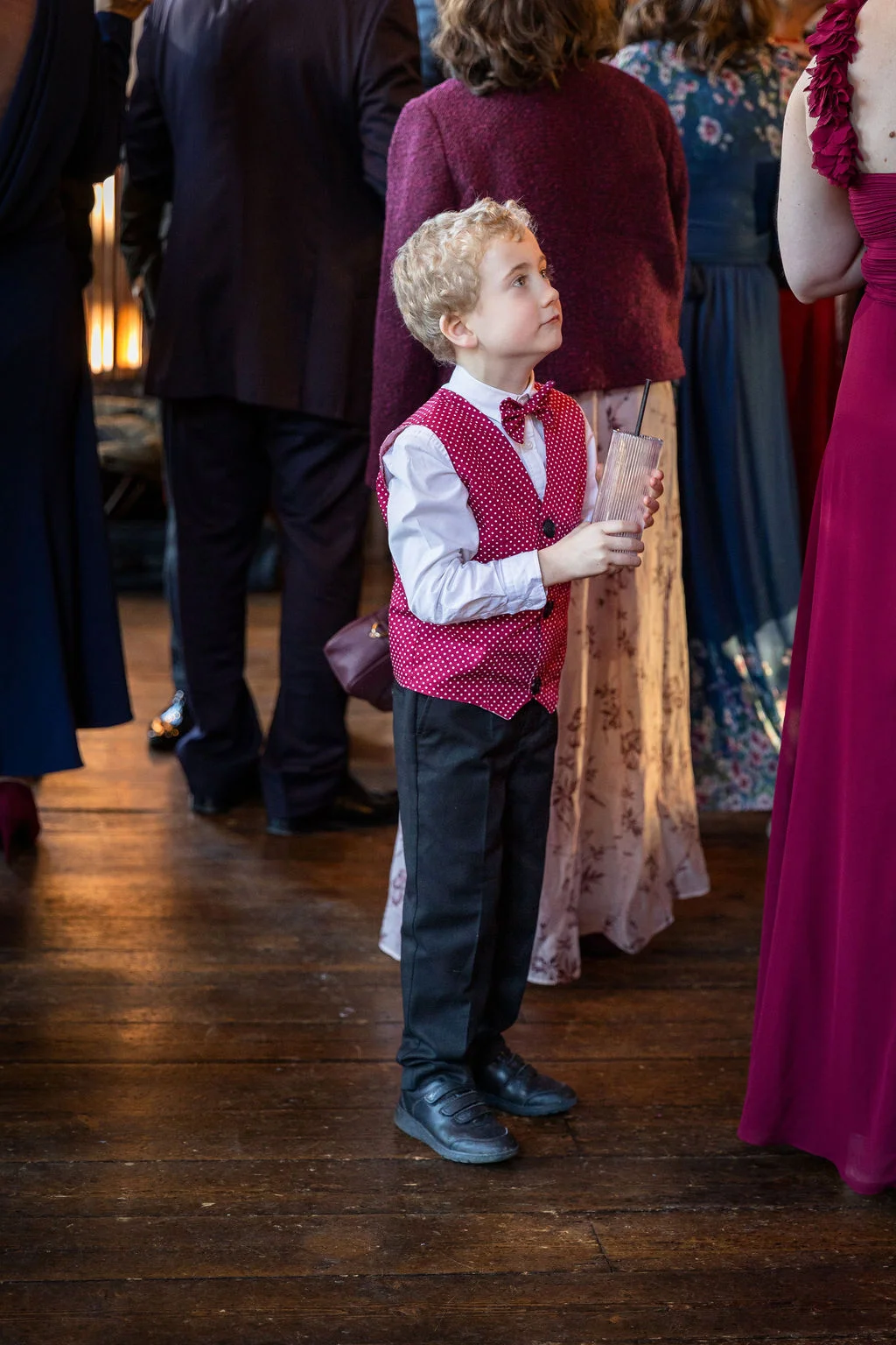 A young page boy wearing a red polka dot waistcoat and matching bow tie stands holding a drink amongst mingling wedding guests inside a rustic barn venue.