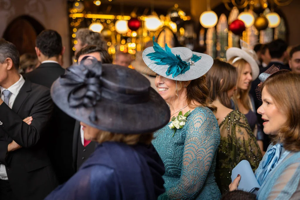 Wedding guests mingling happily at an indoor evening reception, wearing elegant formal hats and fascinators beneath warm fairy lighting.