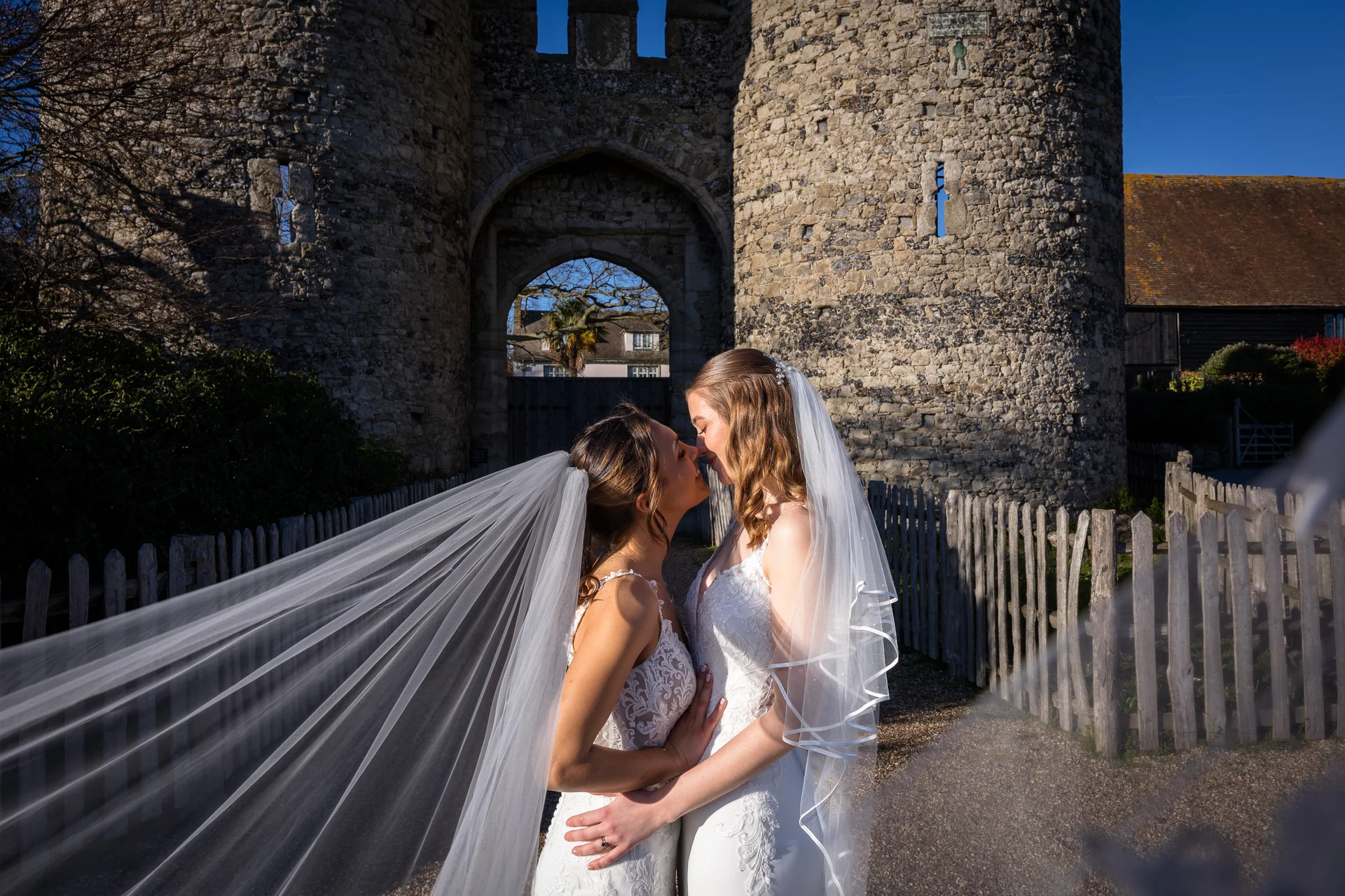 Two brides share a tender kiss in front of an ancient stone castle gate while a long veil billows dramatically in the wind.