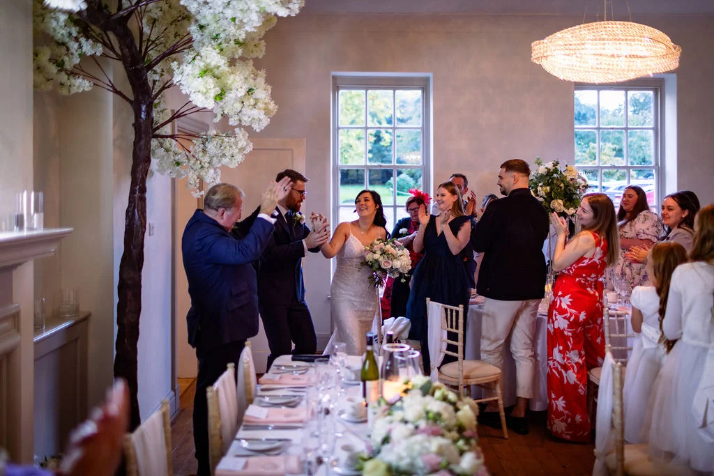 Newlyweds joyfully enter the wedding breakfast reception hand in hand as cheering guests applaud around a beautifully decorated long table.