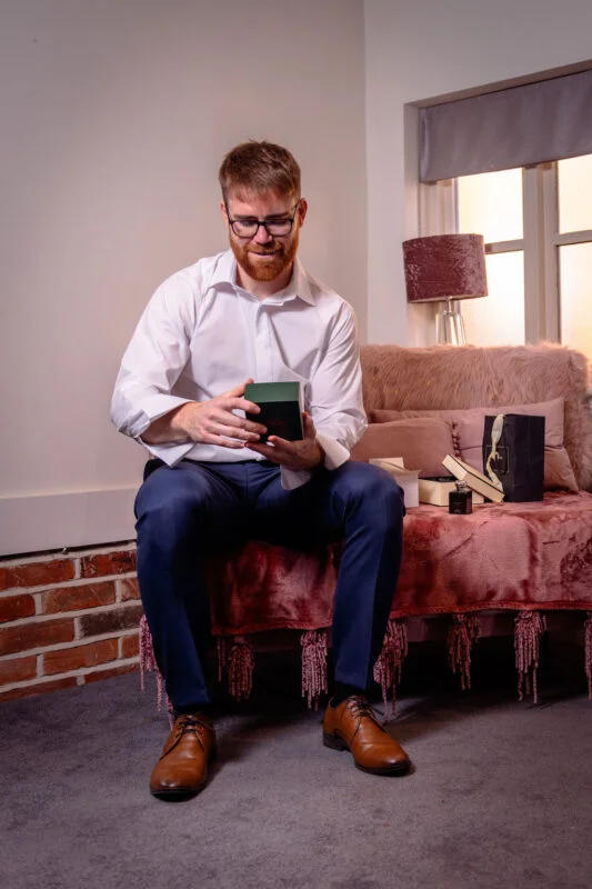 A groom with a ginger beard wearing a crisp white shirt and navy trousers sits on a pink sofa while opening a small green box during wedding preparations.