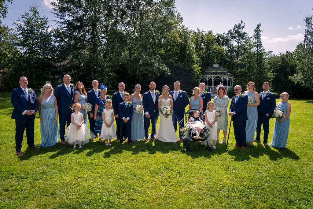 A large wedding party and family group pose together on a sunlit lawn in front of a white gazebo and lush green trees.
