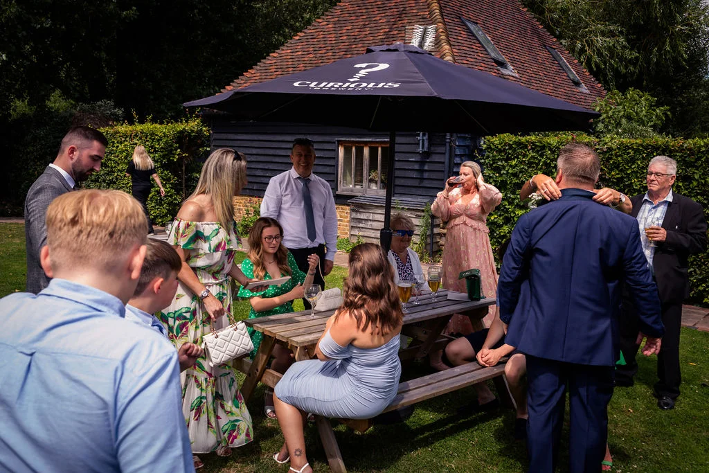 Wedding guests mingle and relax around wooden picnic tables beneath a black parasol in a sunny rustic courtyard.