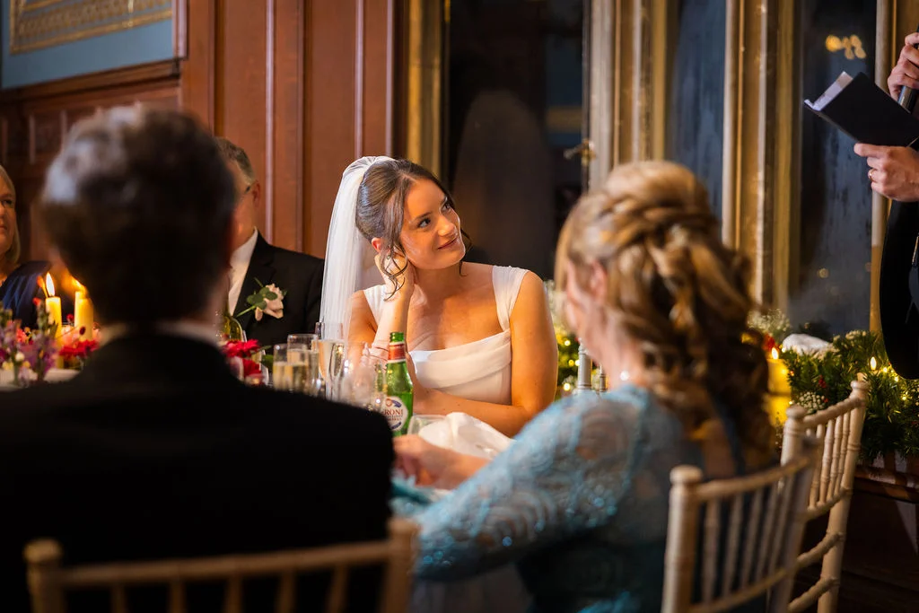 A smiling bride wearing a square neckline dress and veil sits at a candlelit reception table, listening attentively during the wedding speeches.