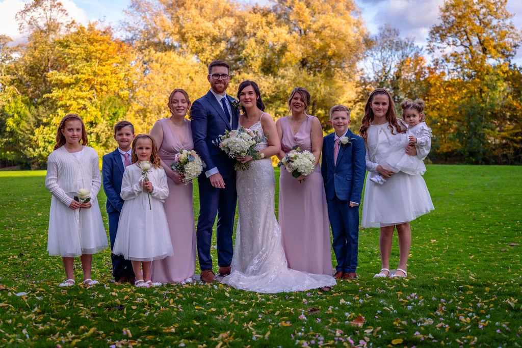 A bridal party including a bride, groom, bridesmaids, and children pose outdoors on a lush green lawn surrounded by vibrant autumn foliage.