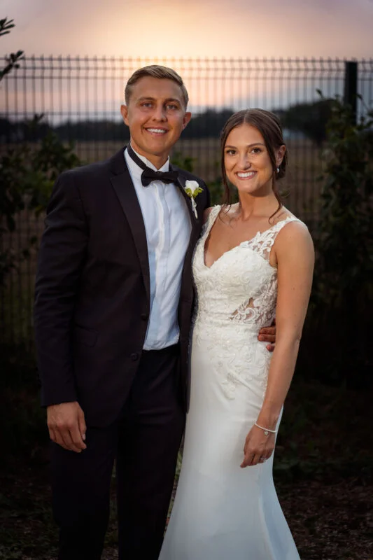 A bride and groom standing in front of a fence at sunset.