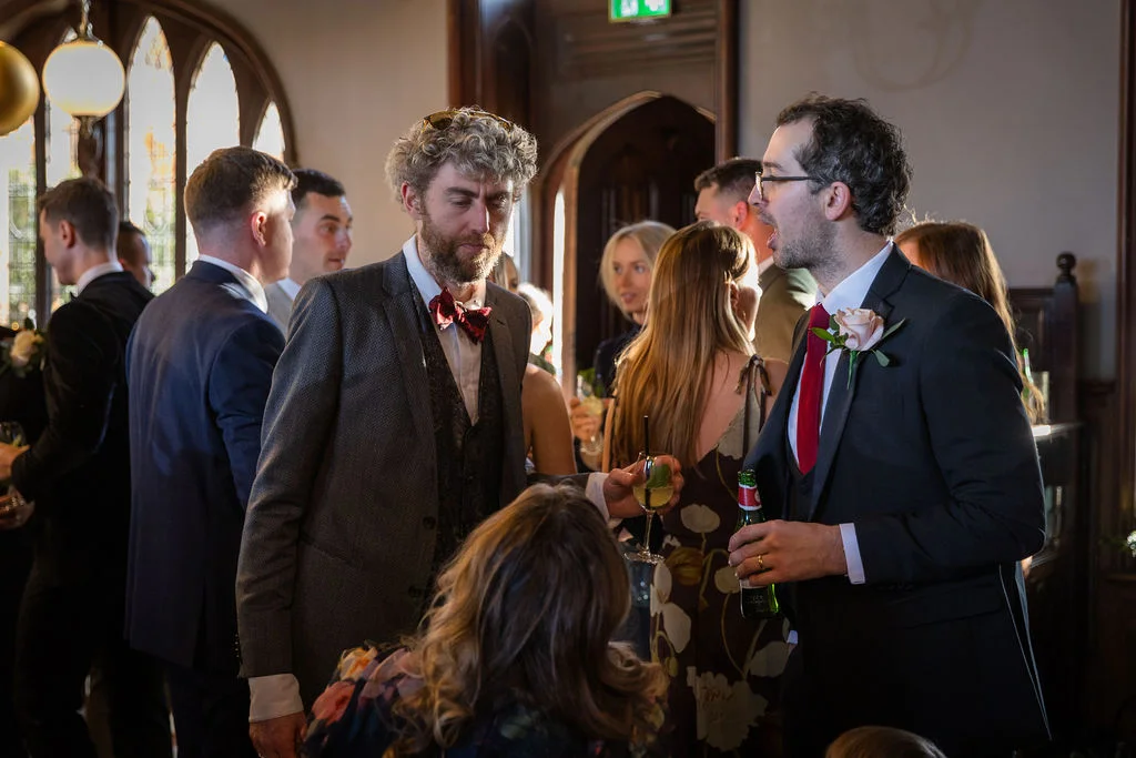 Wedding guests mingle and chat animatedly during the drinks reception in a light-filled historic venue room.