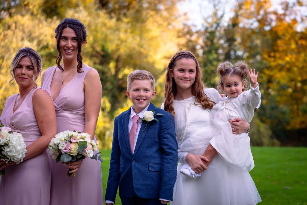 A smiling wedding party including two bridesmaids in pink dresses, a page boy in a blue suit, and young girls in white dresses posing outdoors against a backdrop of golden autumn trees.
