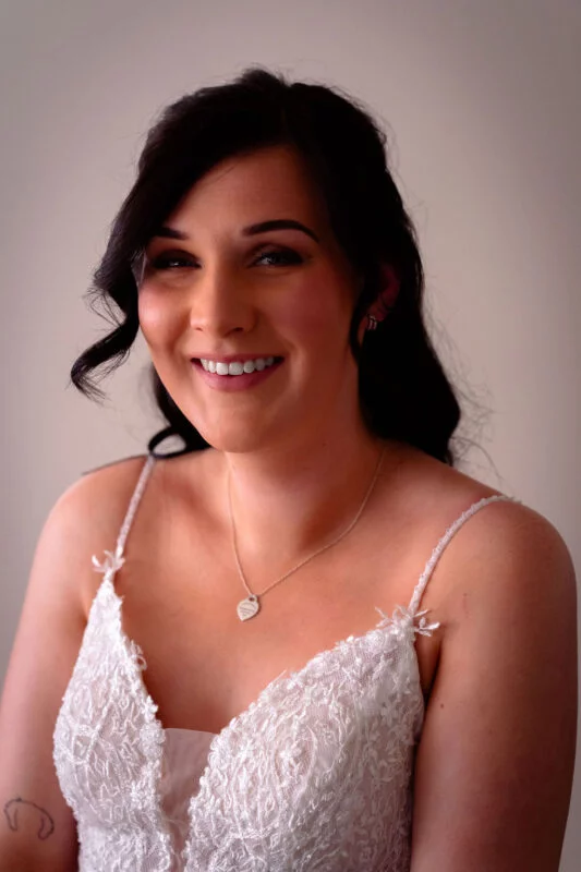 A smiling bride wearing a lace wedding dress with spaghetti straps and a delicate pearl necklace during her bridal portraits.