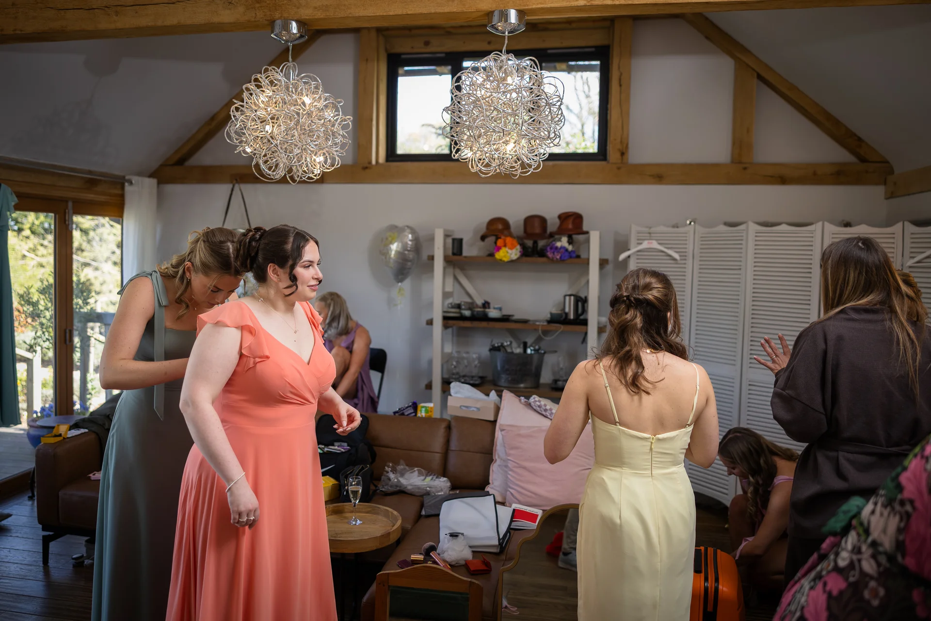 Bridal party members in a rustic barn venue with exposed rafters are getting ready, wearing colourful dresses as final touches are being made.
