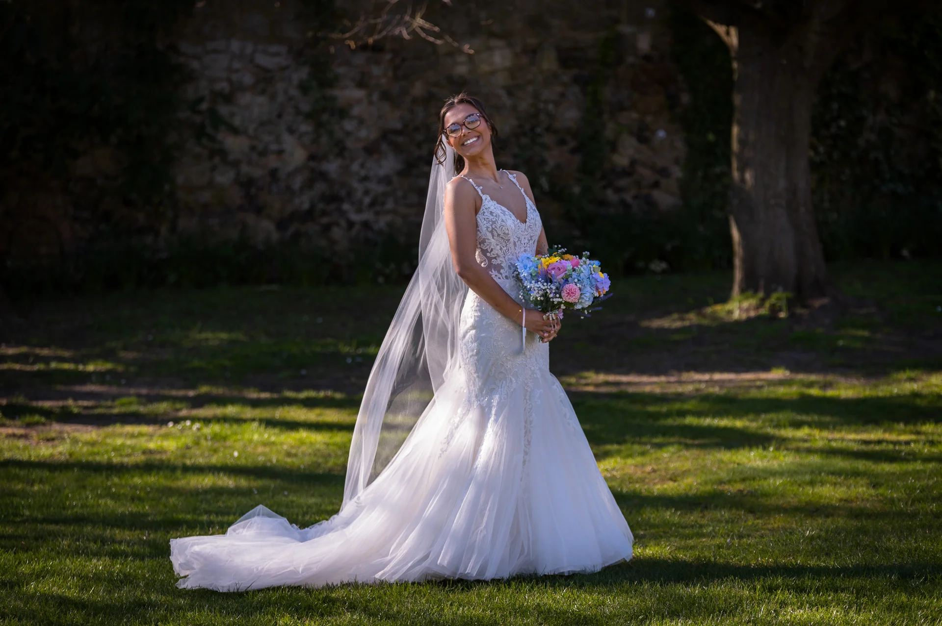 A smiling bride wearing a fit-and-flare lace wedding dress with a sheer corset bodice and a long veil holds a colourful bouquet in a sunny garden.
