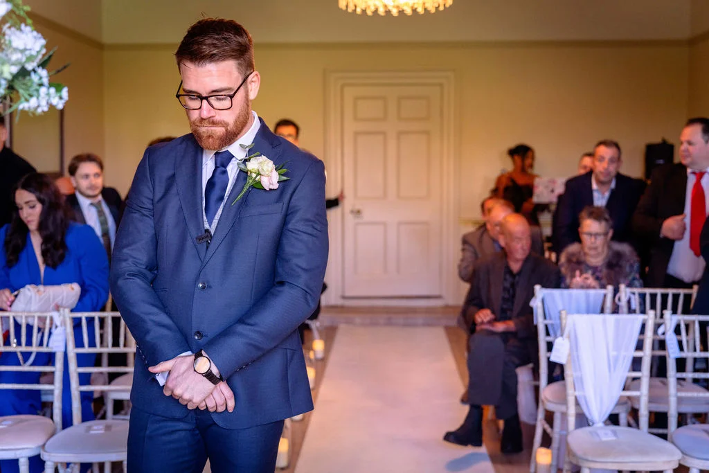 A groom in a sharp navy suit with a boutonniere waits emotionally at the altar while guests look on inside a light-filled venue.