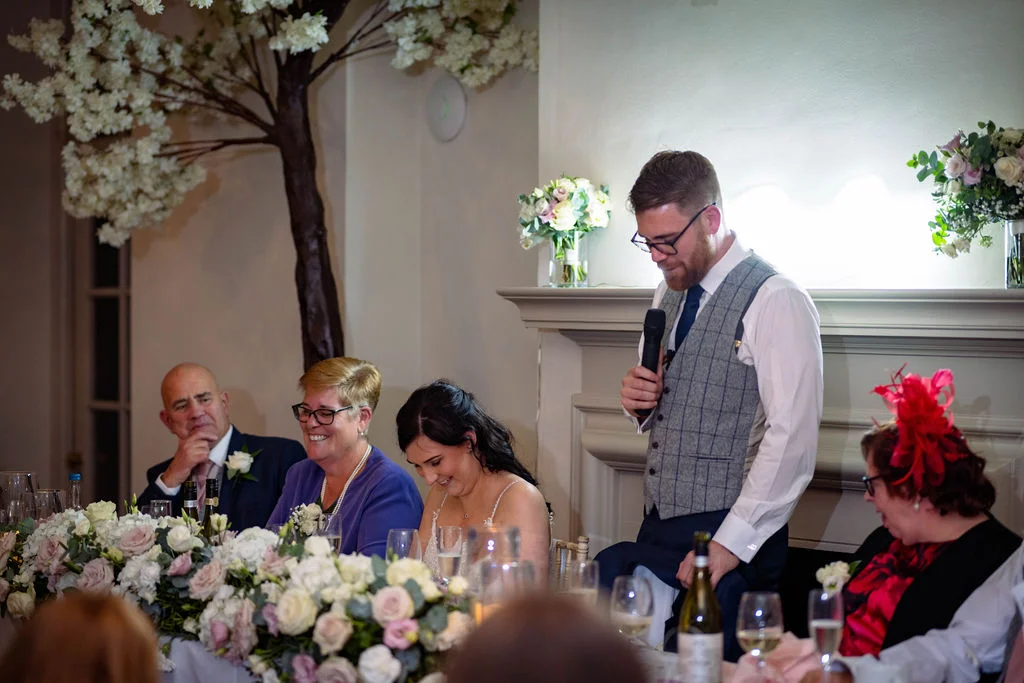 A groom delivers a speech during the wedding reception while seated guests laugh and enjoy the moment in front of a fireplace.