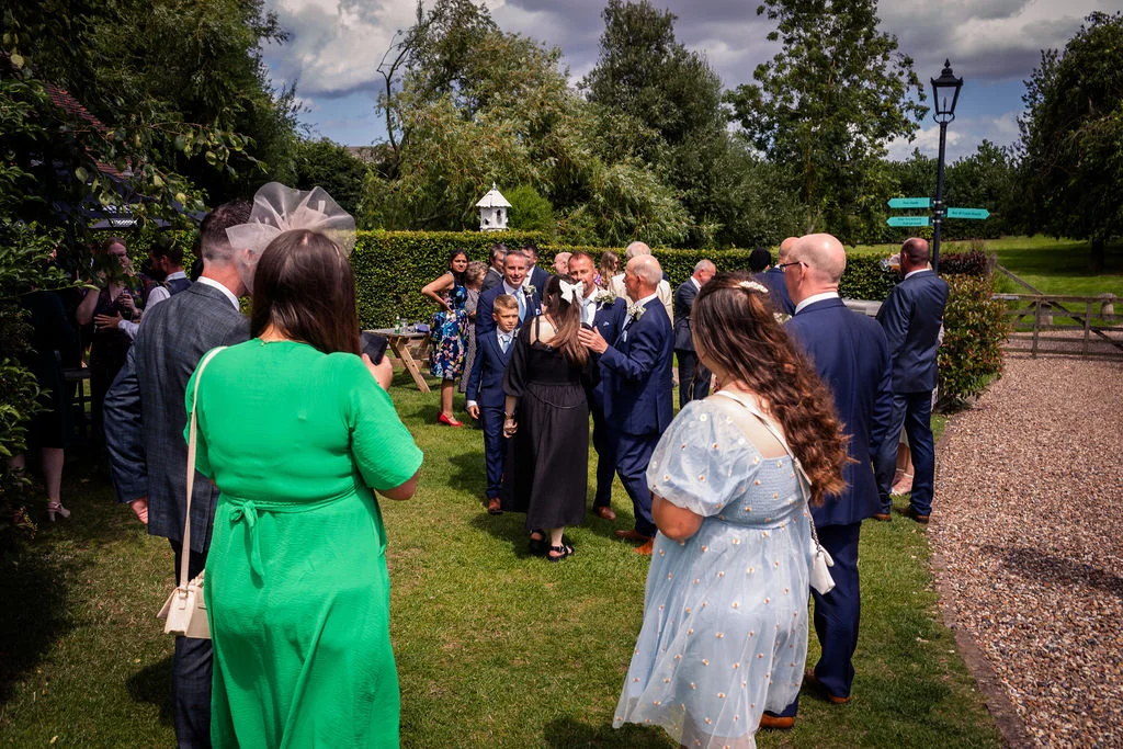 Wedding guests mingle and chat on a sunlit lawn during an outdoor reception surrounded by trees and a tall green hedge.