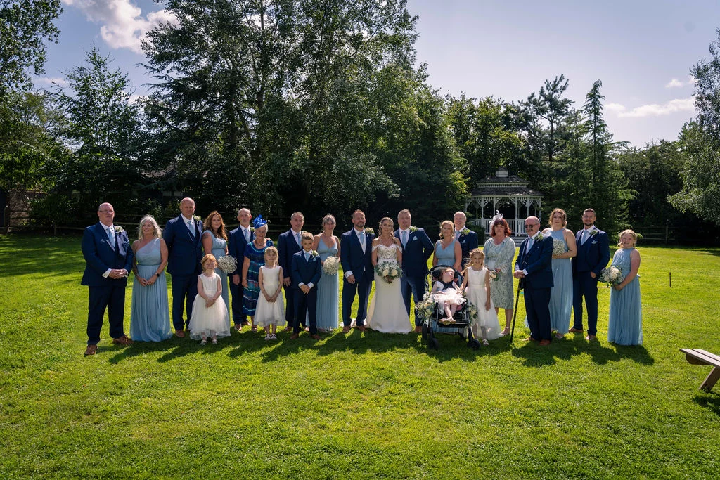 A large wedding party and family group pose for a formal portrait outdoors on a sunny day in a sprawling green garden with mature trees and a gazebo in the background.