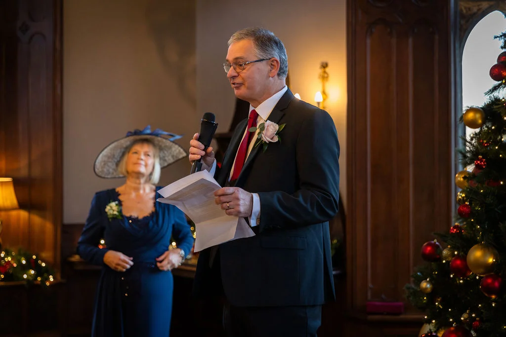 A groom delivers his wedding speech holding a microphone and notes next to a festive Christmas tree while a guest watches in the background.
