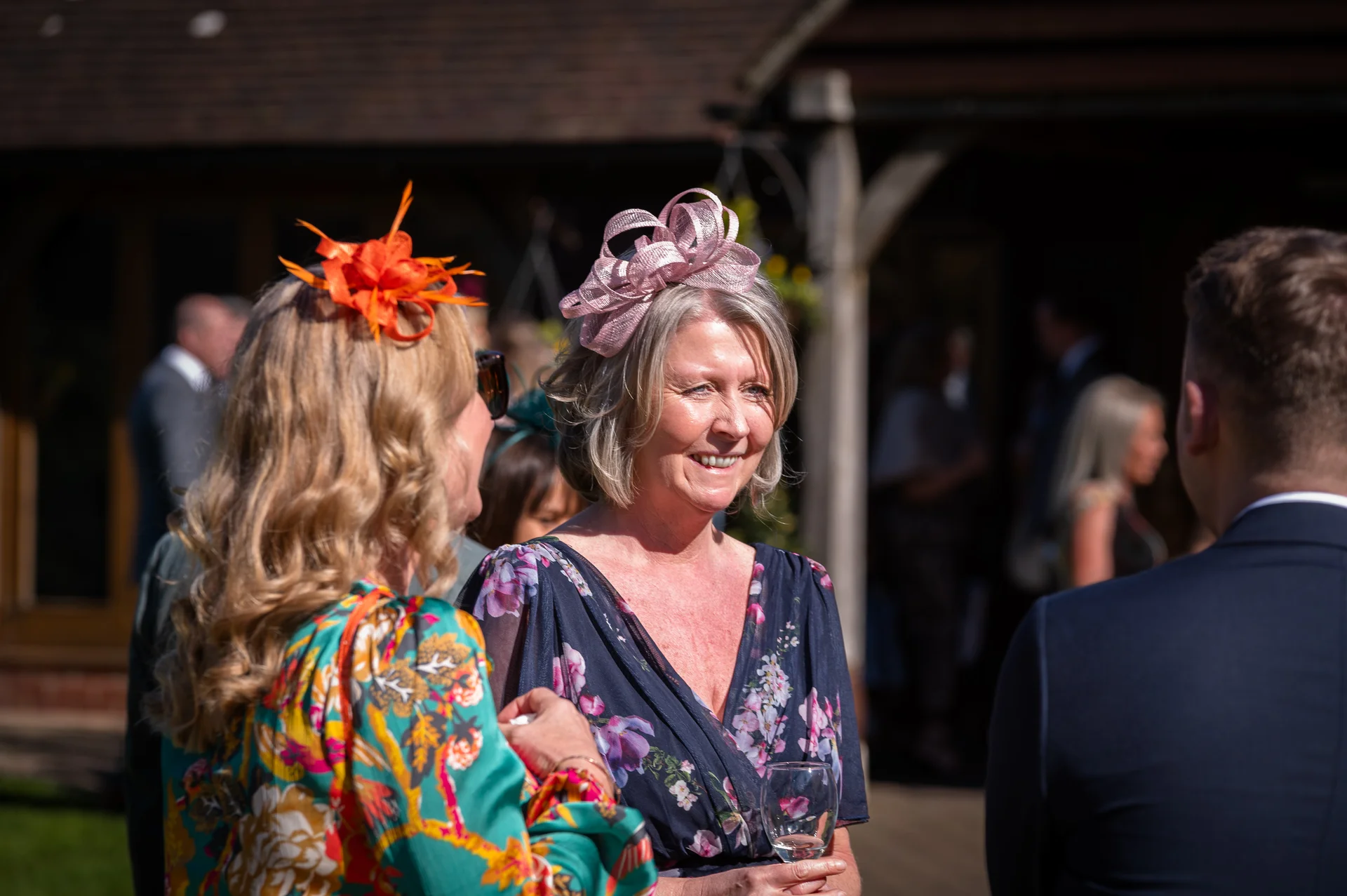 Wedding guests laugh and mingle outdoors in the sunshine during the drinks reception, wearing colourful dresses and elegant fascinators.
