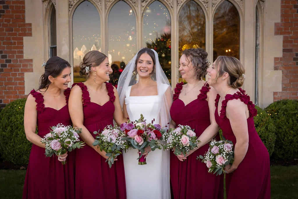 A joyful bride in a white square-neck gown stands smiling alongside four bridesmaids wearing matching burgundy dresses holding floral bouquets outside a historic brick building.