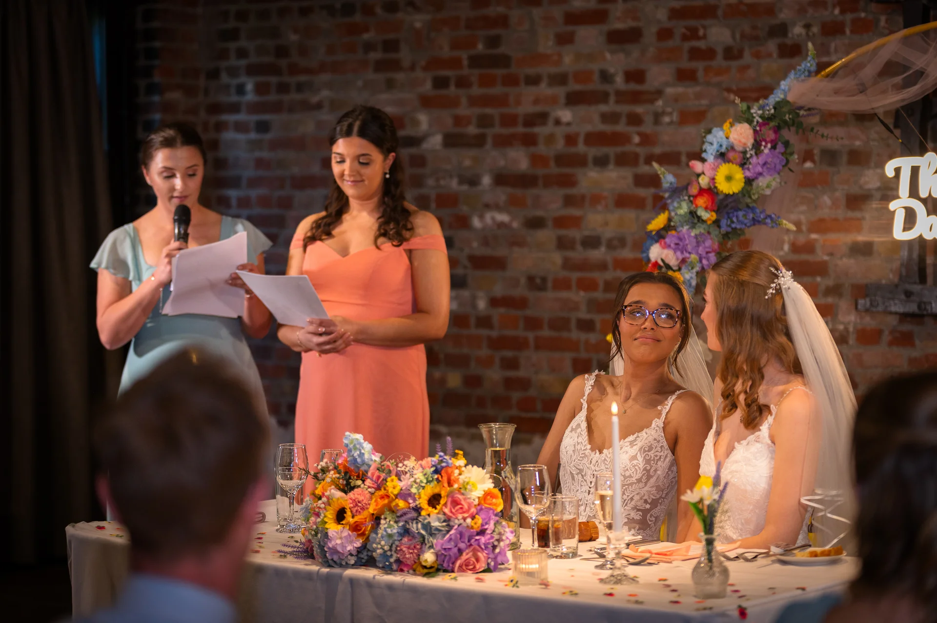 Two brides sit at the top table listening to a speech, with two bridesmaids standing and reading from notes in front of an exposed brick wall.