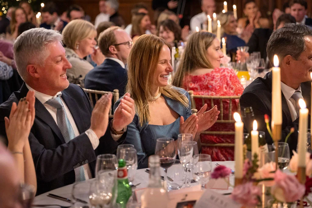 Wedding guests sit around a candlelit round table during the wedding breakfast, laughing and applauding warmly during the speeches.