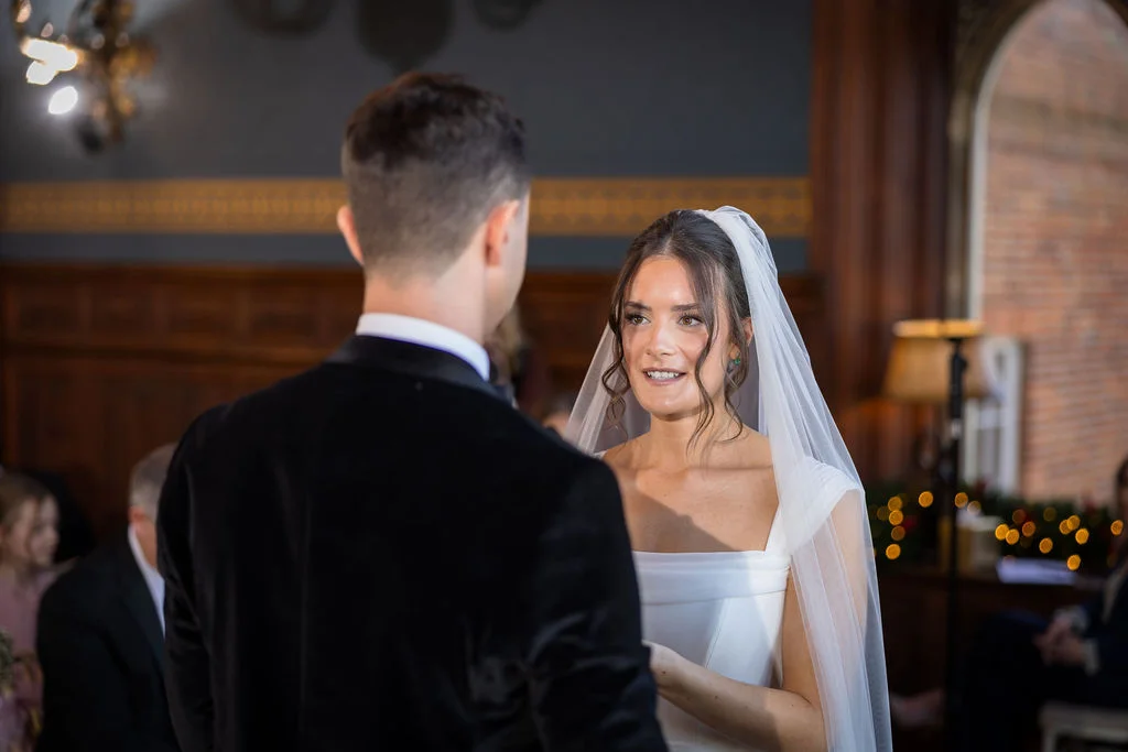 A groom in a velvet tuxedo faces his bride during their wedding ceremony inside a wood-panelled room, with the bride wearing a square neckline gown and chapel veil.
