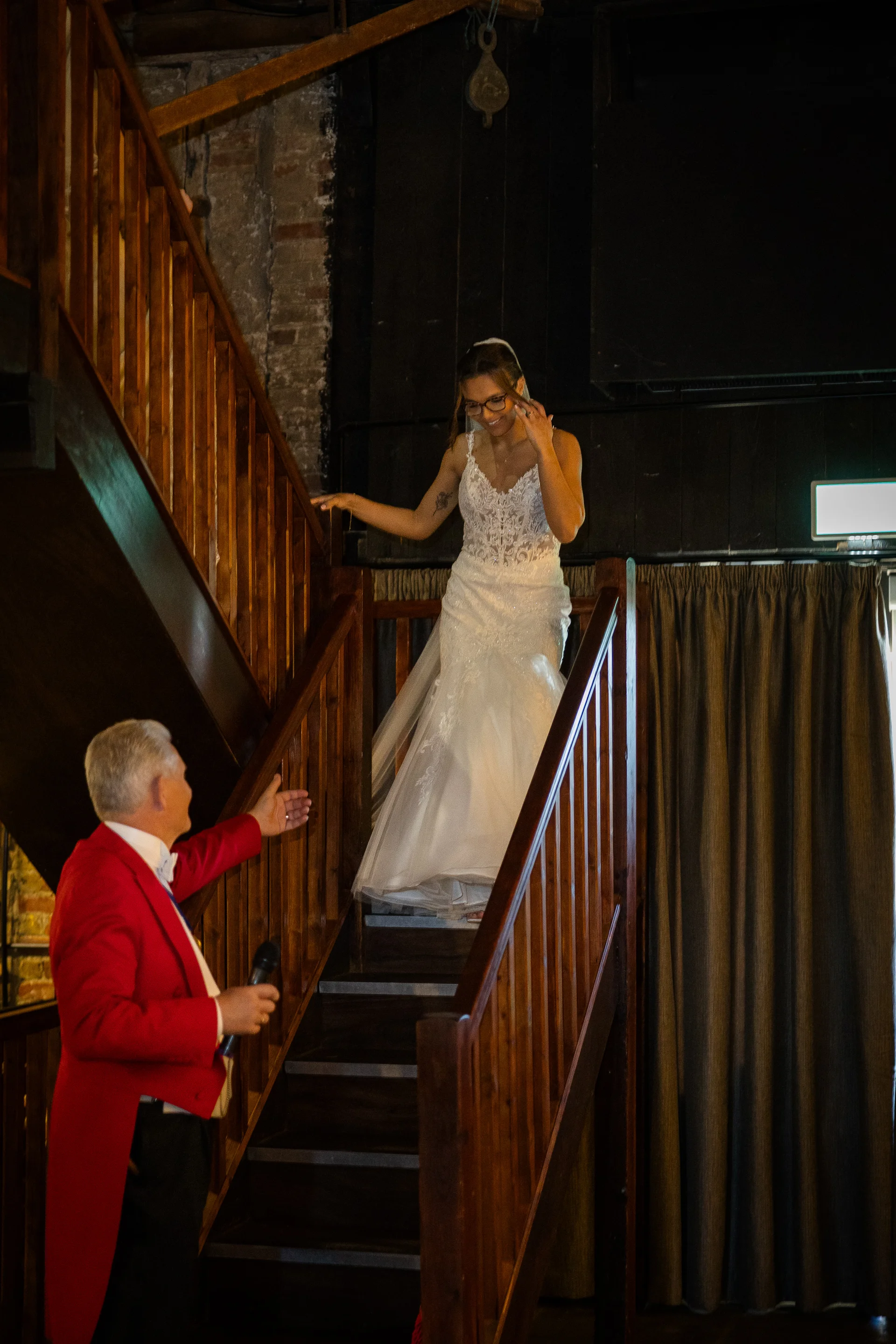 A smiling bride wearing a lace wedding dress descends a wooden staircase inside a rustic venue while a toastmaster in a red tailcoat gestures towards her.