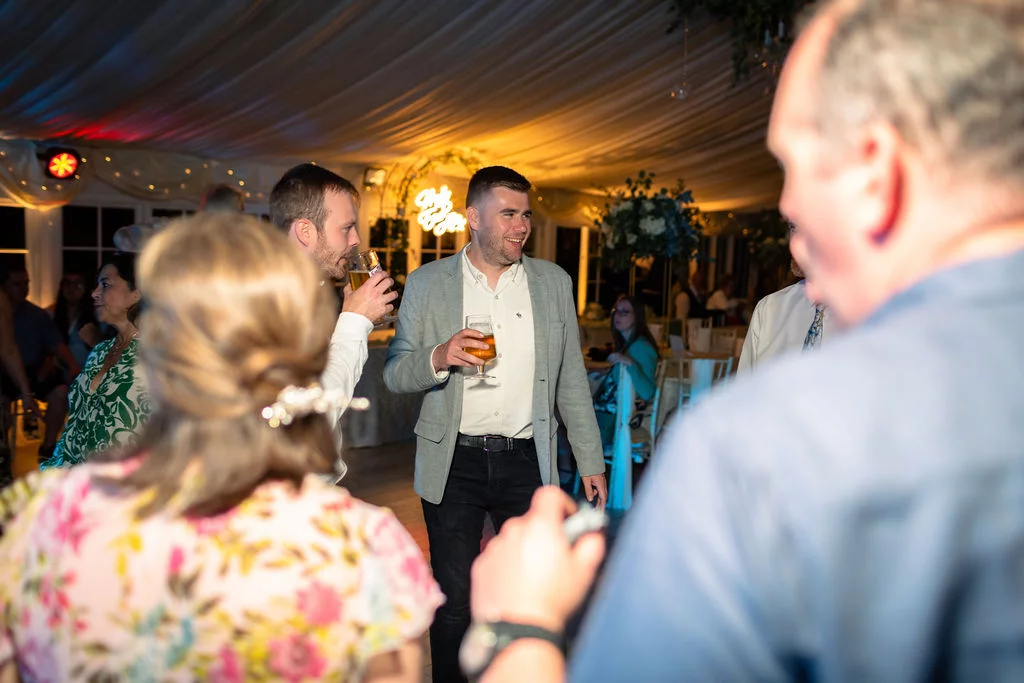 Wedding guests mingle and enjoy drinks inside a marquee with draped white ceiling fabric during the evening reception.