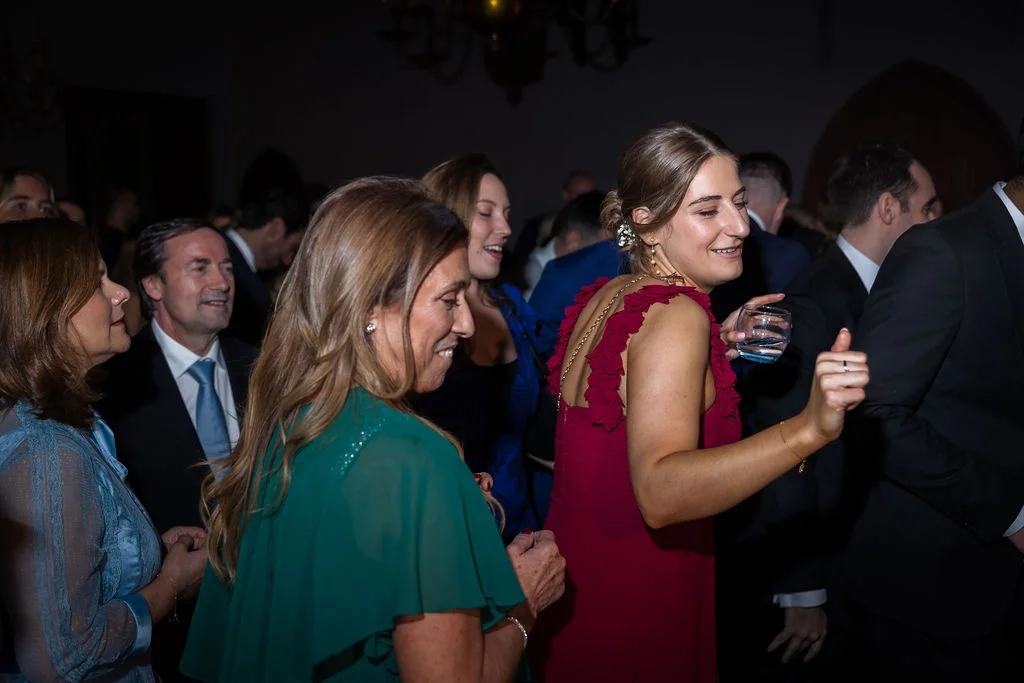 Wedding guests enjoy the evening reception, with a woman in a red ruffled dress dancing and holding a glass amidst a lively crowd.