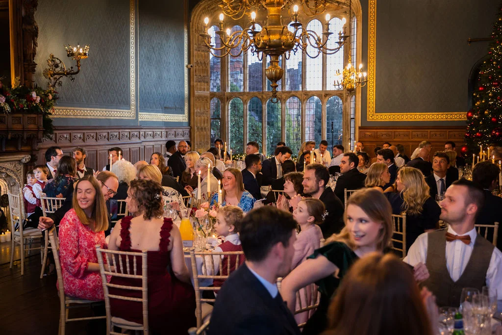 Wedding guests enjoy a festive wedding breakfast inside a grand, wood-panelled stately home with large arched windows and a glittering Christmas tree.