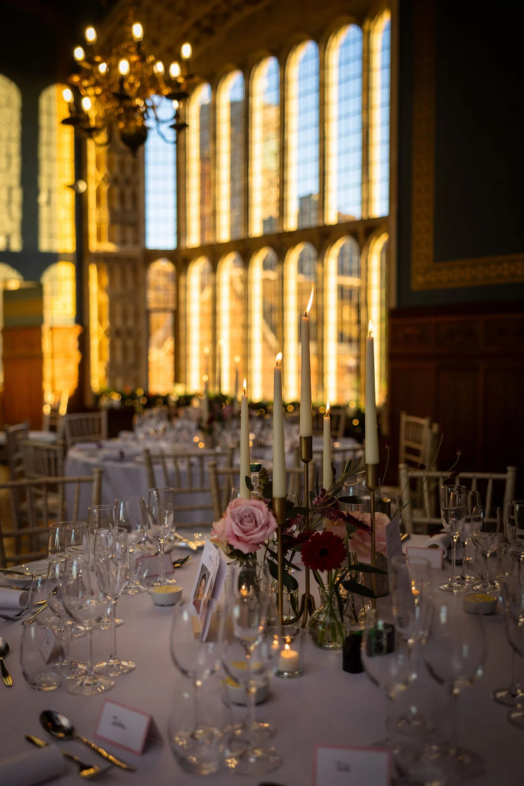 An elegant wedding reception table features flickering taper candles, a delicate floral arrangement with pink roses, and crystal glassware inside a grand hall with towering arched windows.