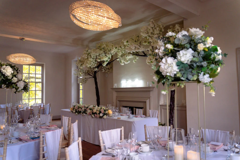 An elegantly decorated wedding reception room featuring tall floral centrepieces on gold stands, white tablescapes, and a stunning white blossom arch over the top table.