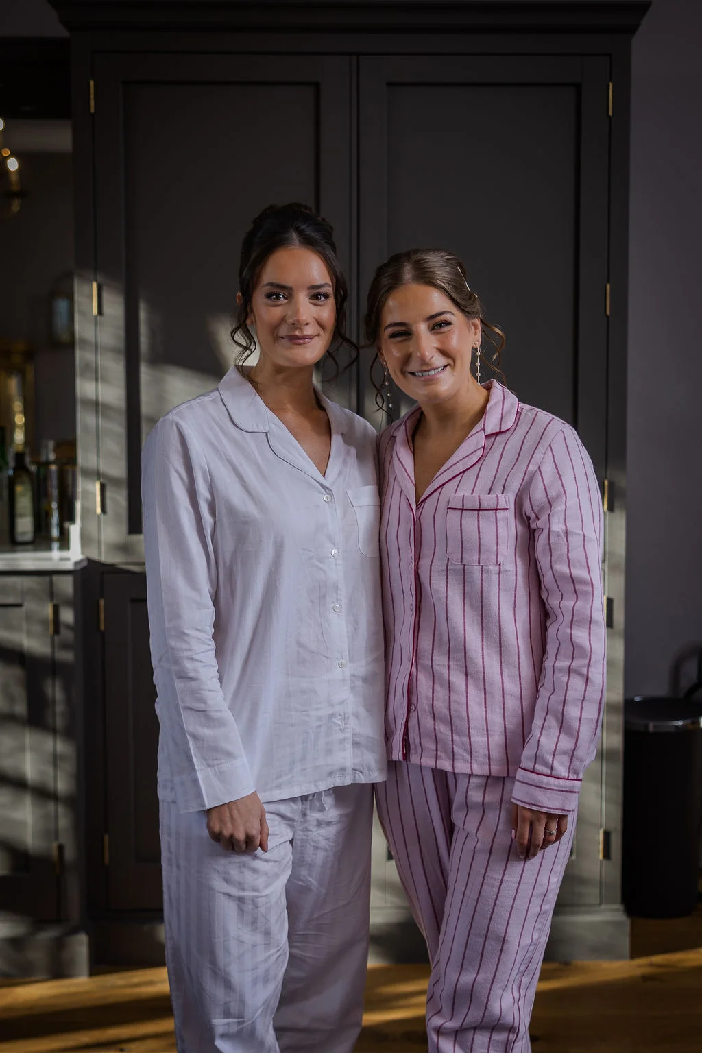 Two smiling brides stand side by side indoors during morning preparations, wearing matching pyjamas.