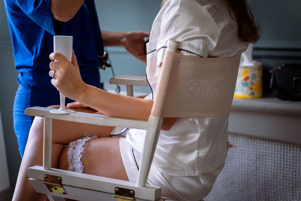A bride in matching silk pyjamas sits in a director chair holding a glass while getting ready, wearing a white lace garter.