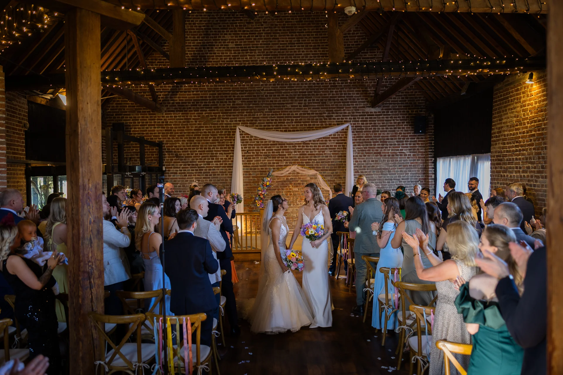 Two brides walk hand in hand down the aisle of a rustic brick barn venue, smiling at each other as cheering guests applaud and toss petals.