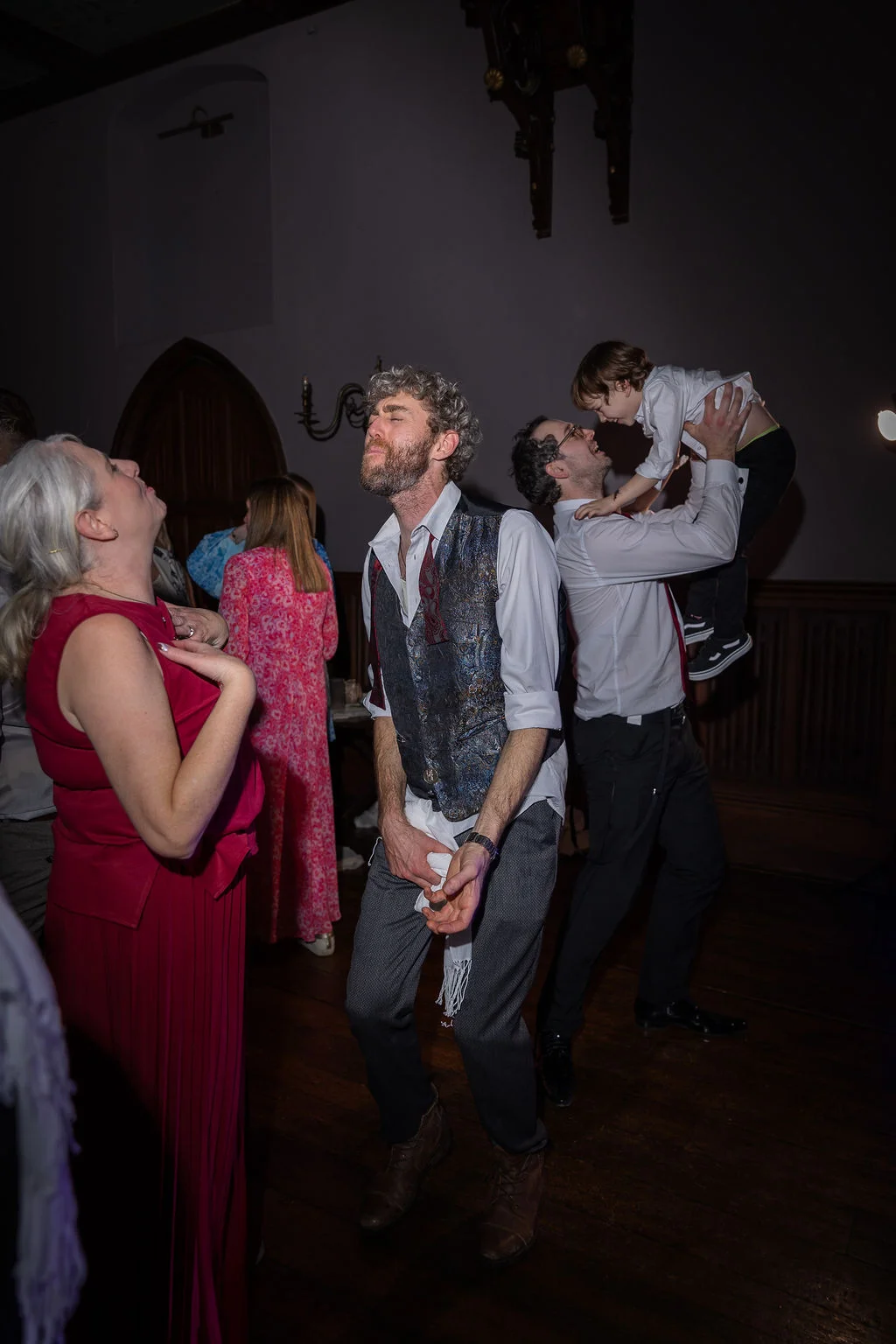 Wedding guests enjoy the evening reception with a man lifting a child into the air while others dance and laugh together.