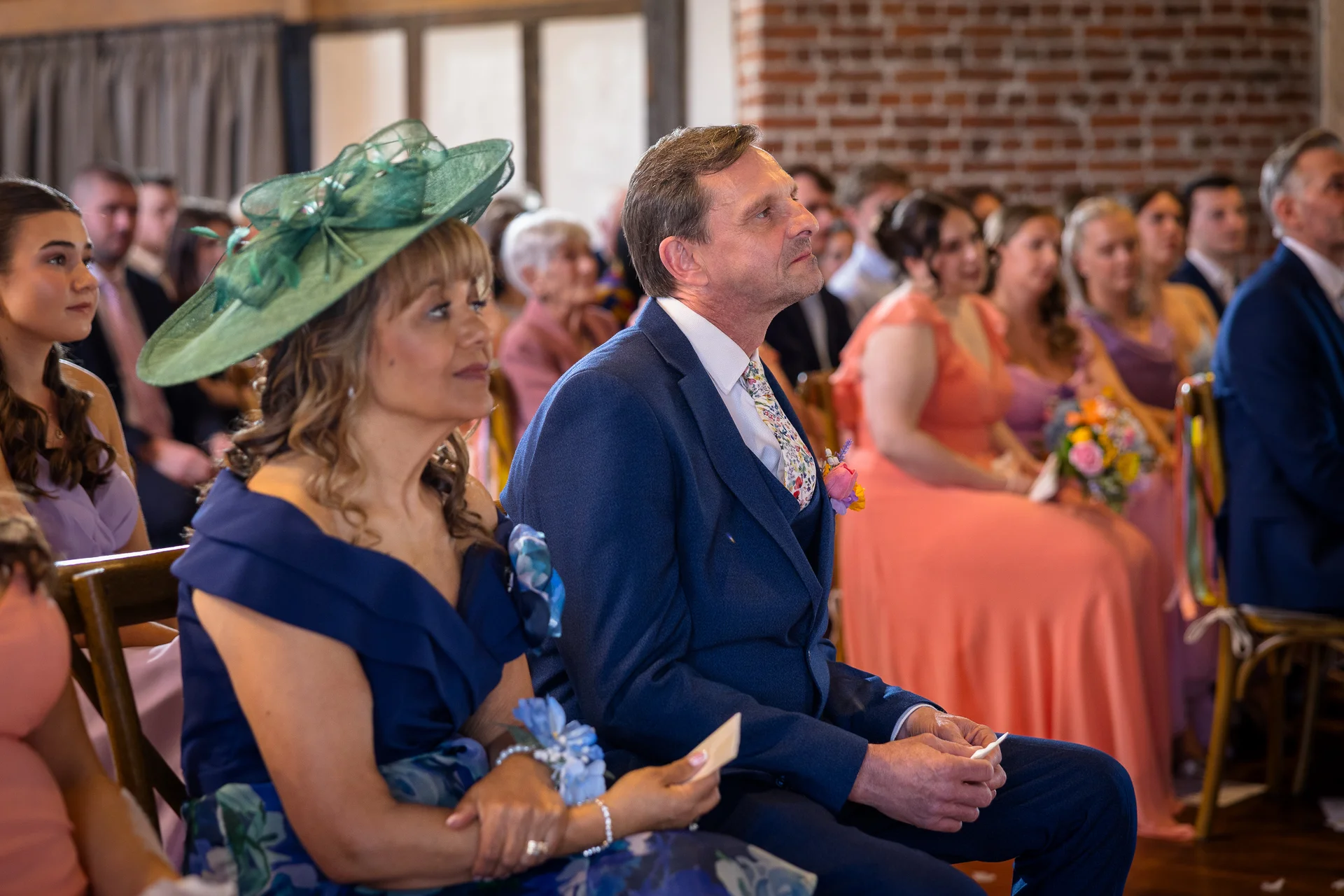 A mother and father of the bride seated attentively among wedding guests during a ceremony inside a venue with exposed brick walls.
