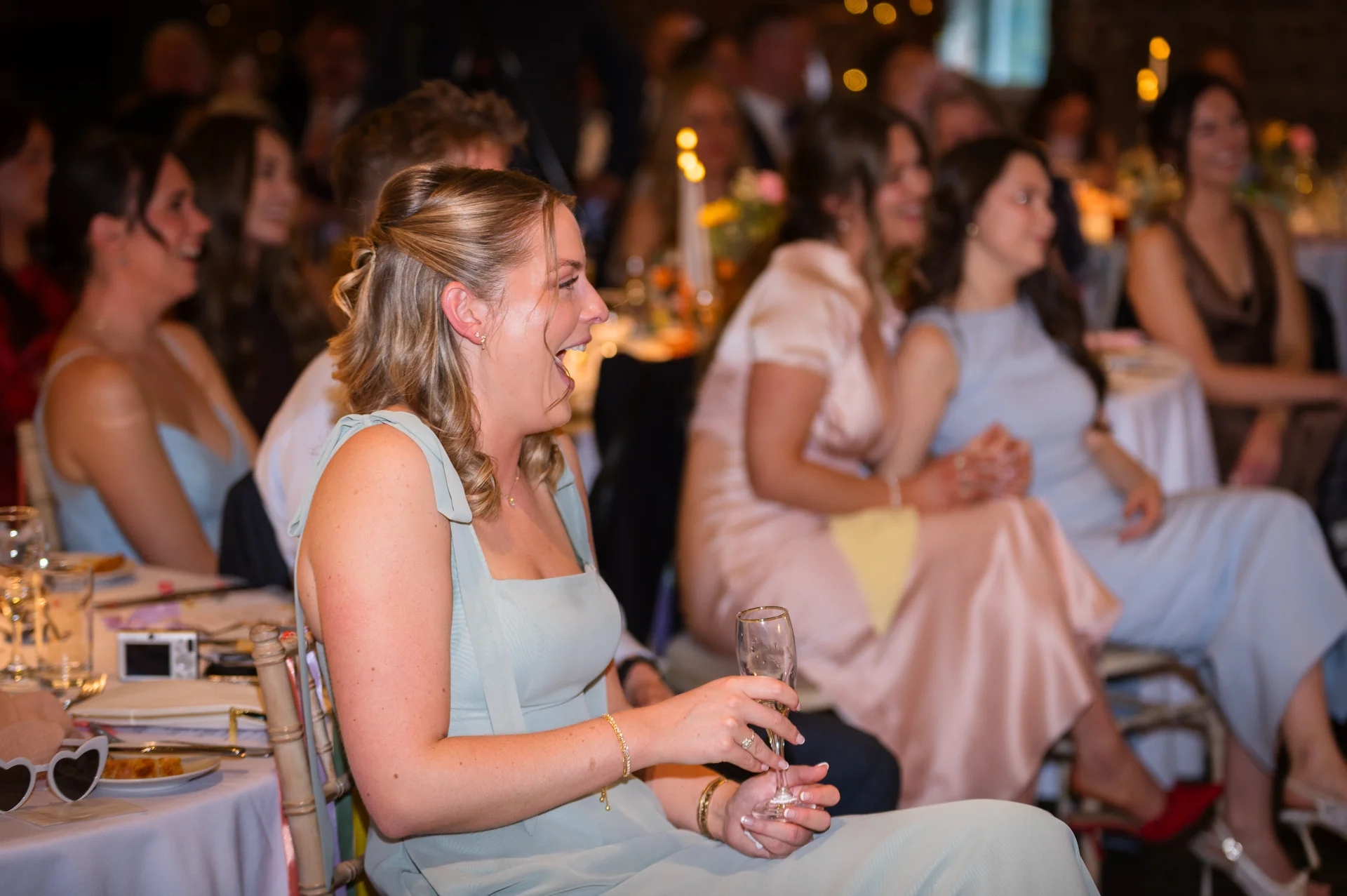 A guest in a sage green dress laughs heartily while holding a glass of champagne at a wedding reception table.