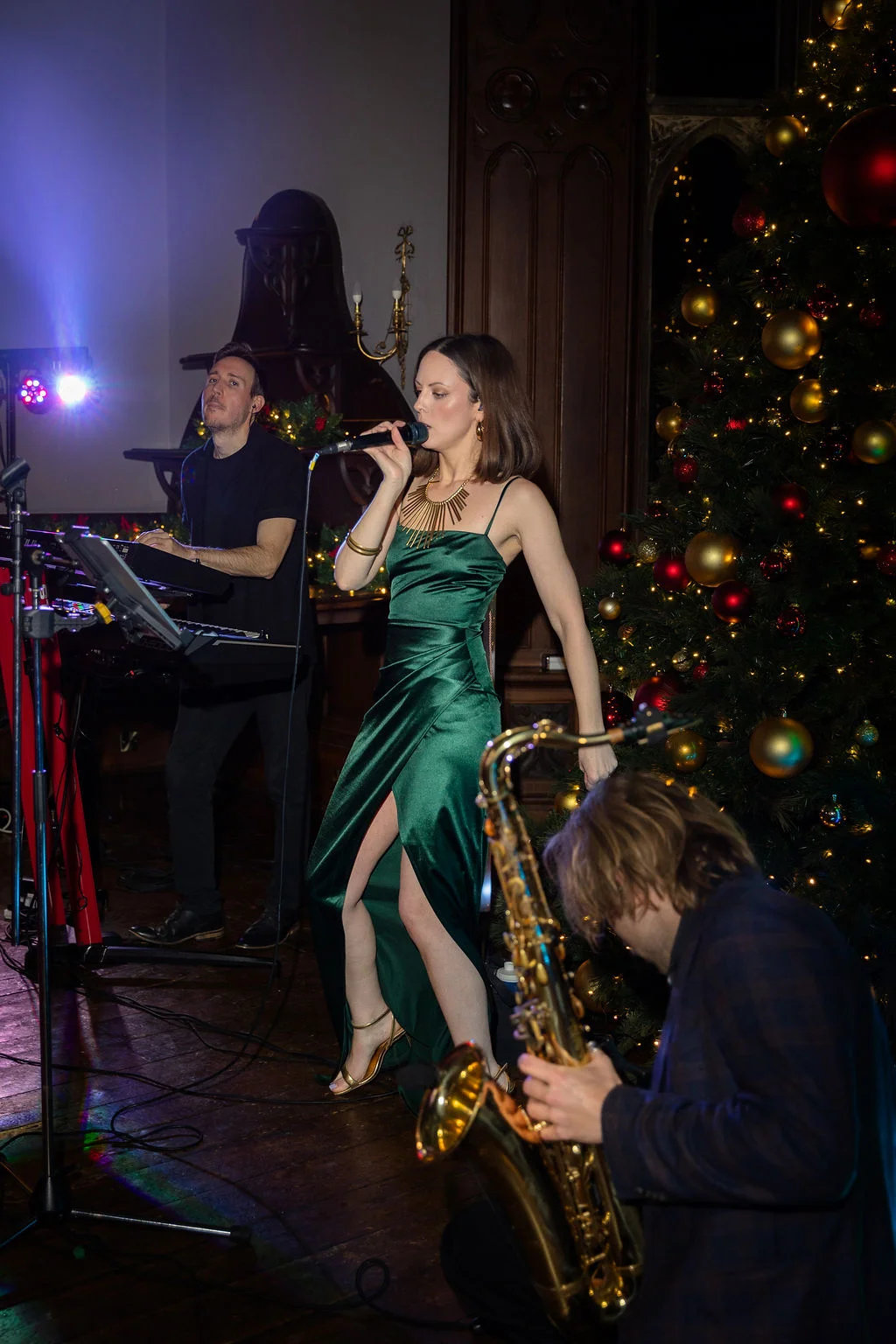 A live wedding band performs at a festive evening reception near a large illuminated Christmas tree, featuring a female singer in a green dress and a saxophonist.