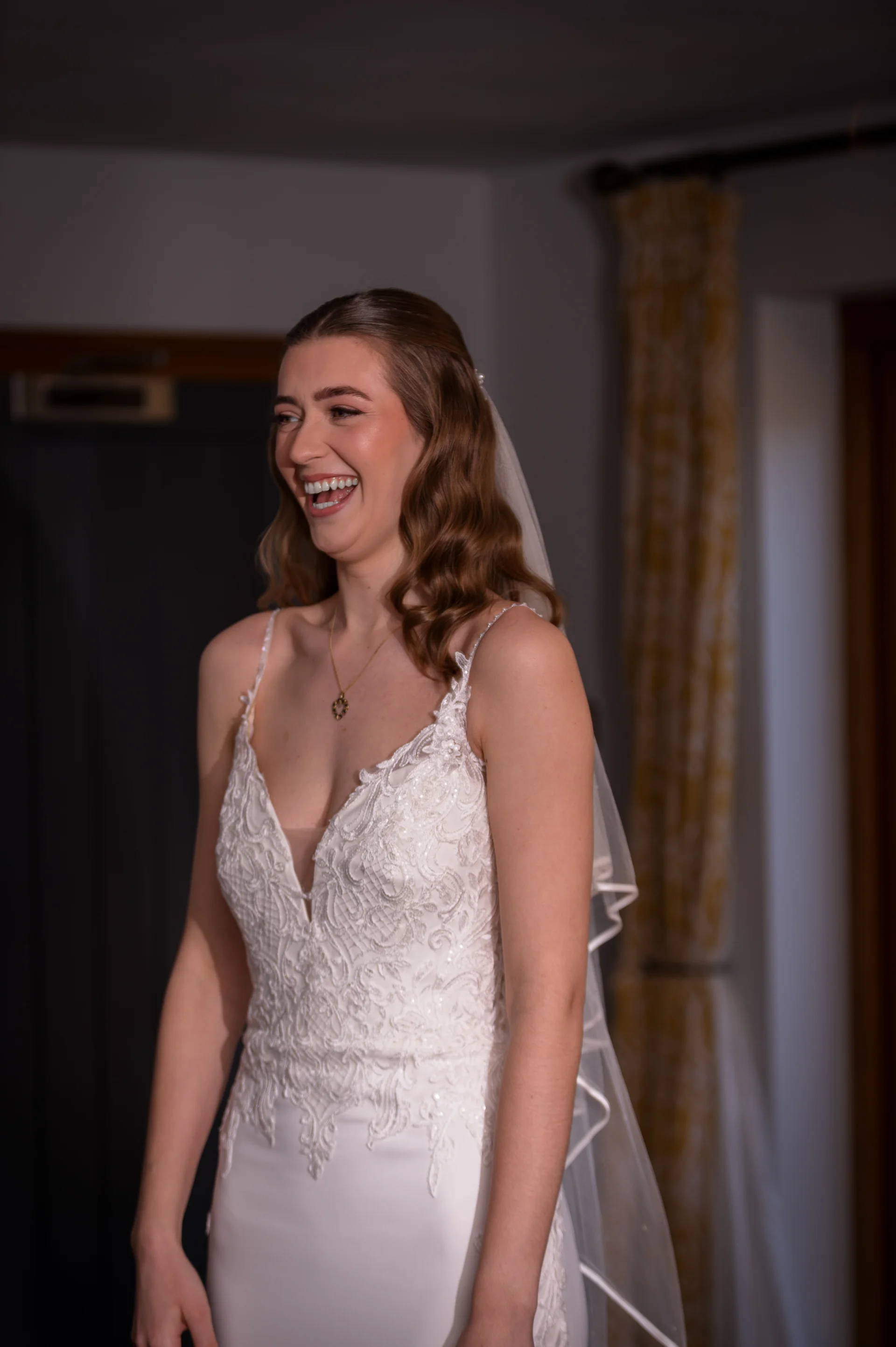 A smiling bride with half-up wavy hair wearing a lace v-neck wedding dress and veil laughs happily indoors.