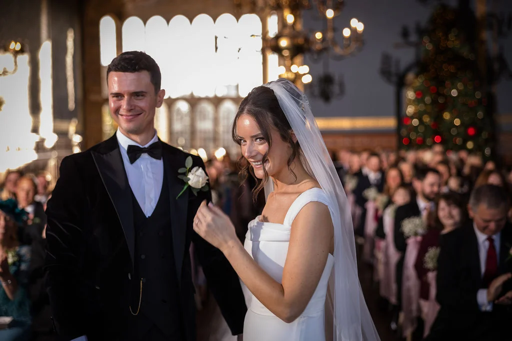 A newlywed couple smiles at the camera during their ceremony inside a grand stately home with guests seated in the background.