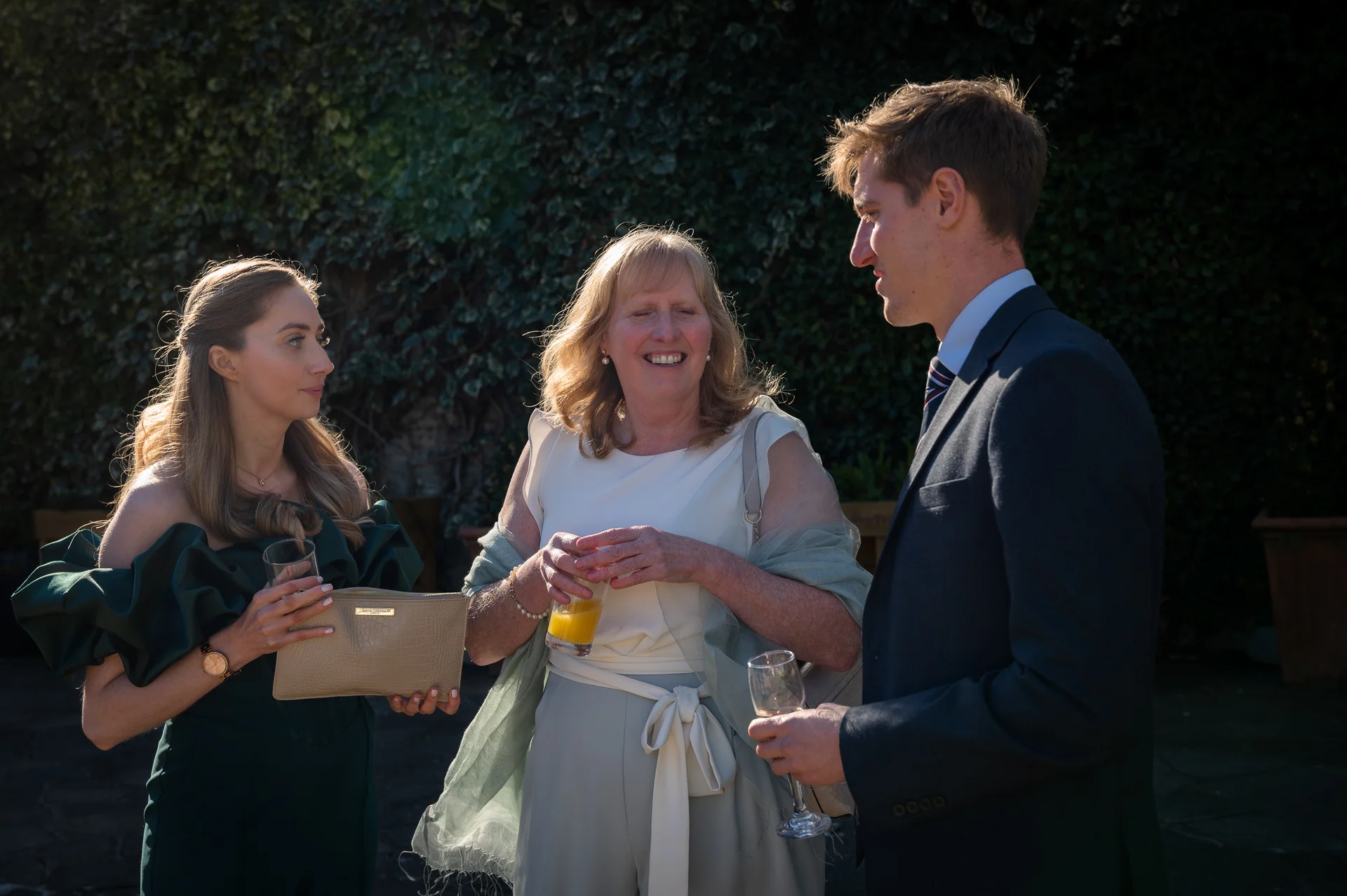Three wedding guests chat and share a drink outdoors in the warm sunlight.