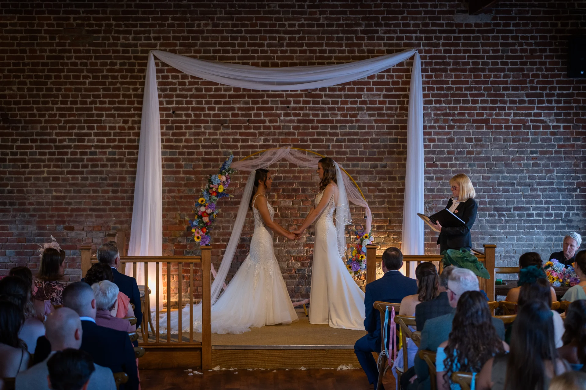 Two brides hold hands during their wedding ceremony in front of an exposed brick wall decorated with a fabric arch and floral arrangements.