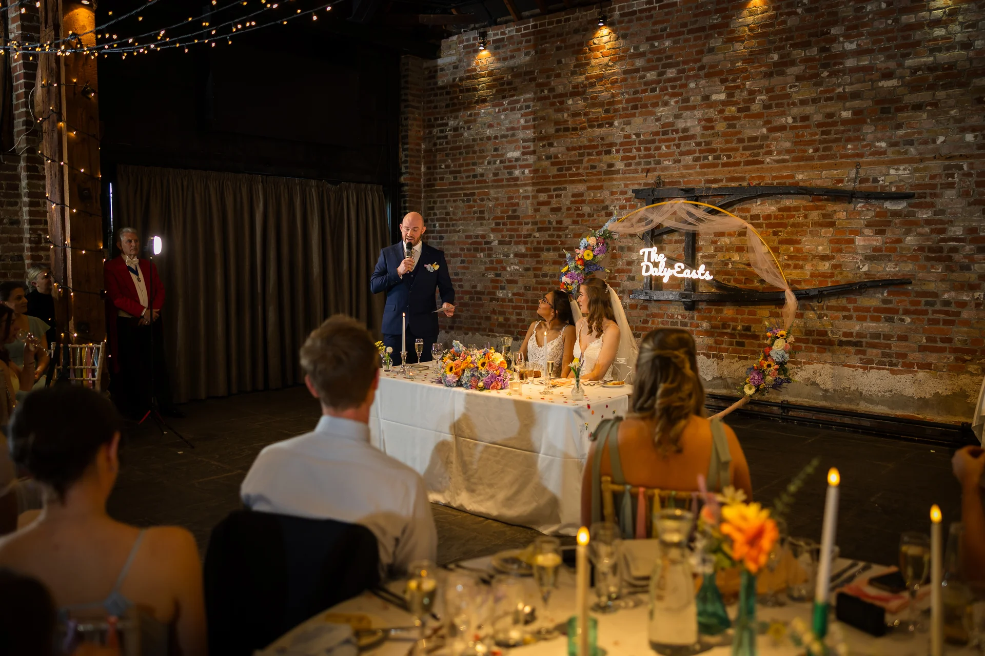 A groom gives a speech at the top table during a wedding reception in an industrial warehouse venue featuring exposed brick walls and a custom neon sign.
