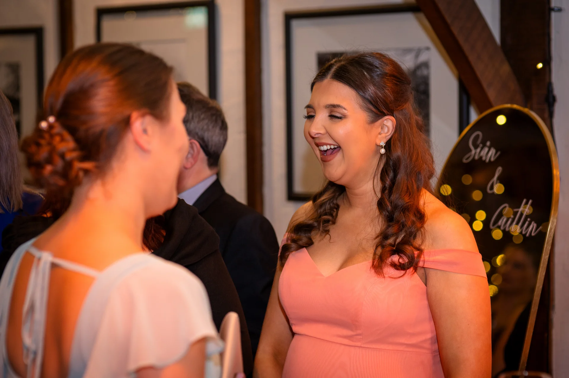 A guest wearing an off-the-shoulder pink dress laughs heartily while mingling at a wedding reception near a welcome mirror.