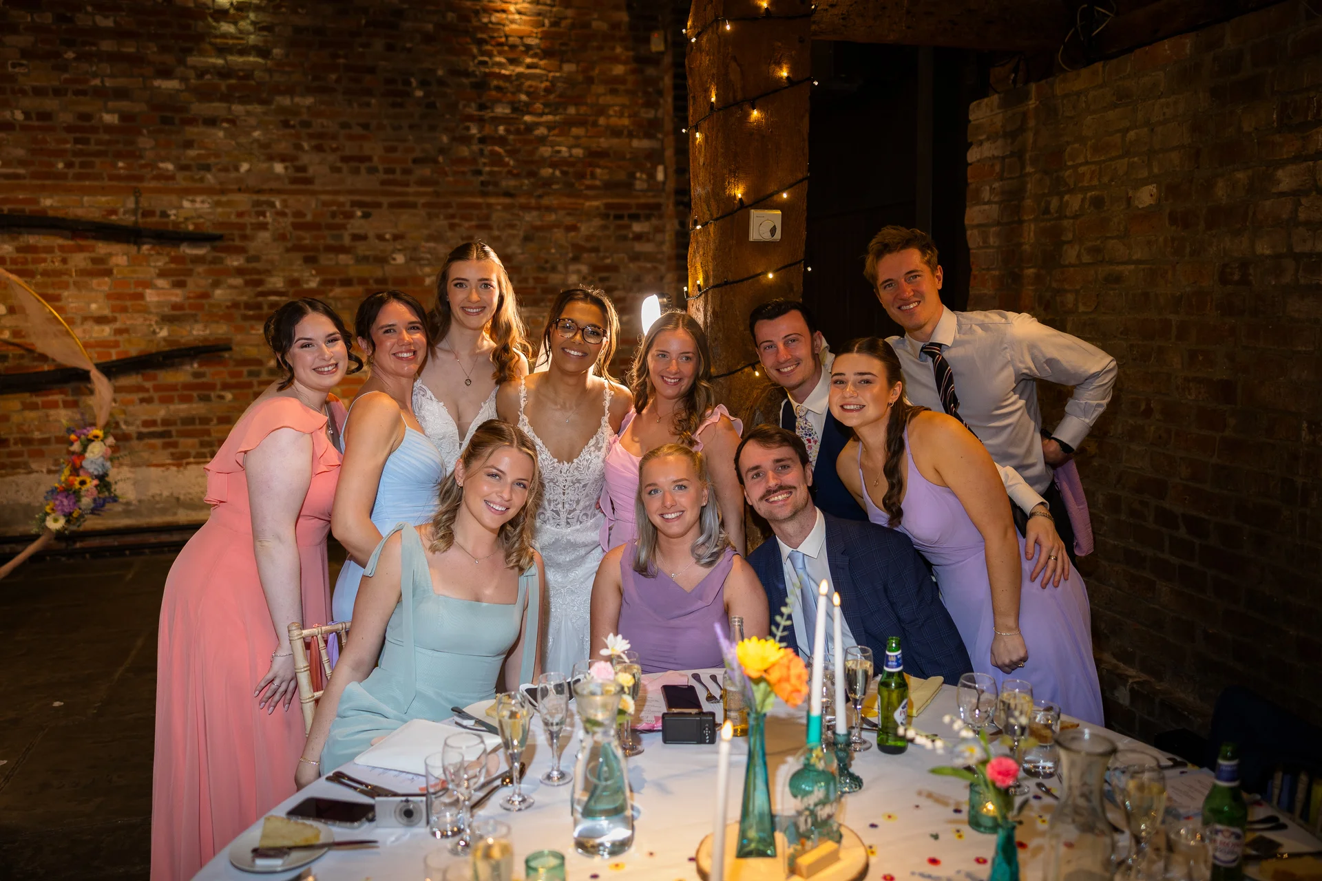 A cheerful wedding group poses together around a candlelit banquet table inside a rustic brick barn venue adorned with twinkling lights.