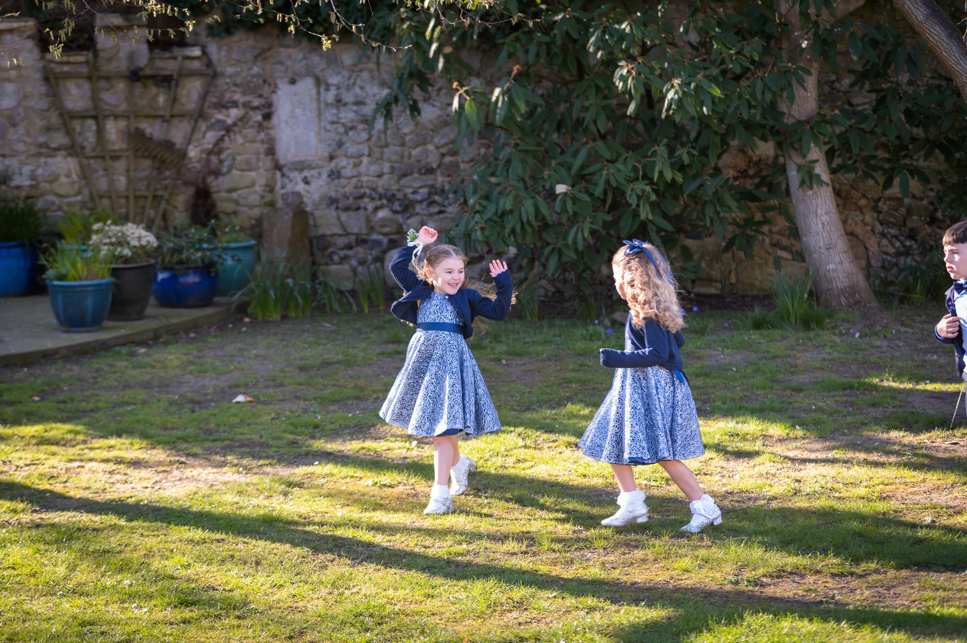 Two young flower girls in patterned blue dresses and matching cardigans joyfully dance and play on a sunlit lawn in front of a rustic stone wall.