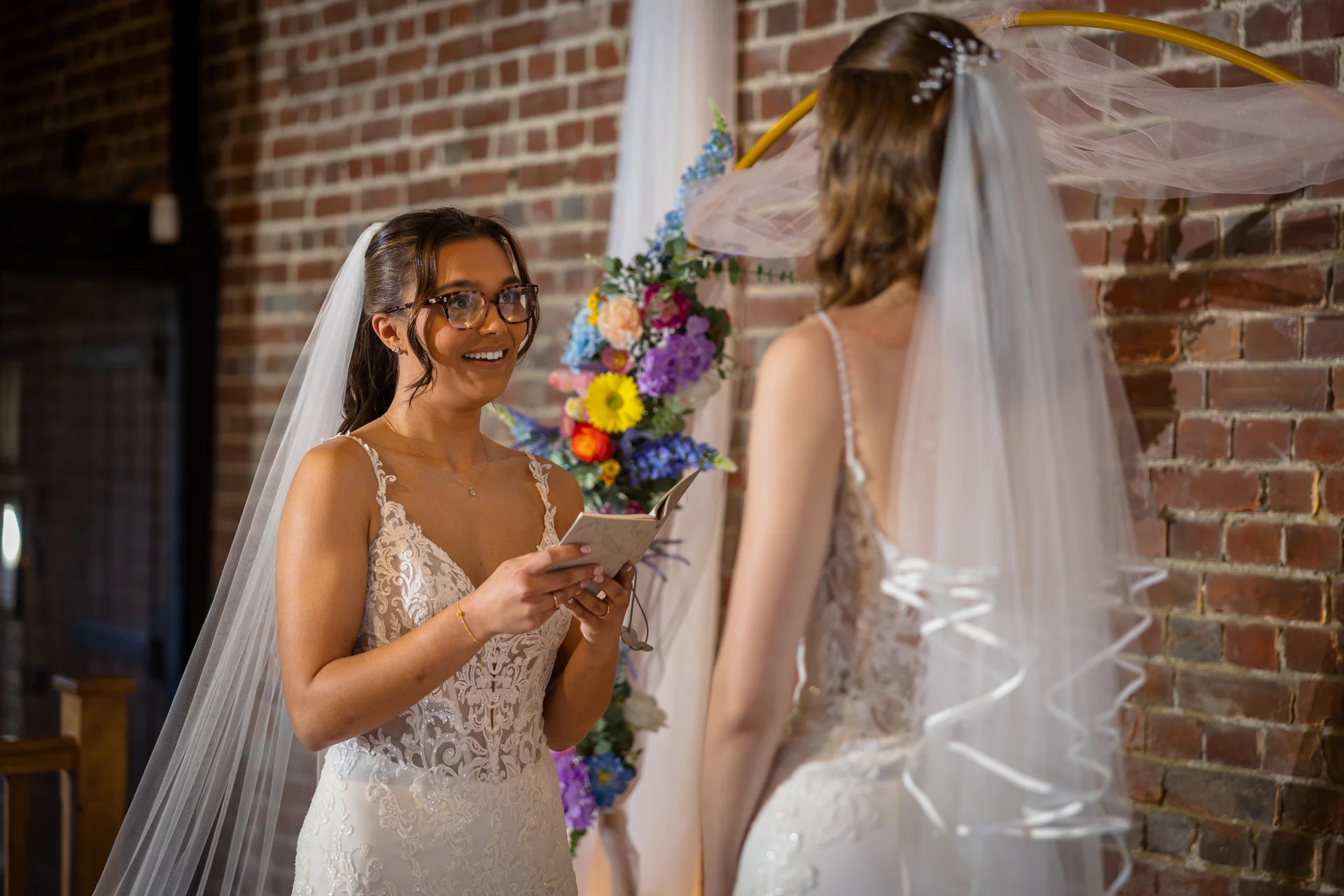 Two smiling brides stand opposite each other in front of a brick wall and floral arch, with one reading personal vows from a booklet during their wedding ceremony.