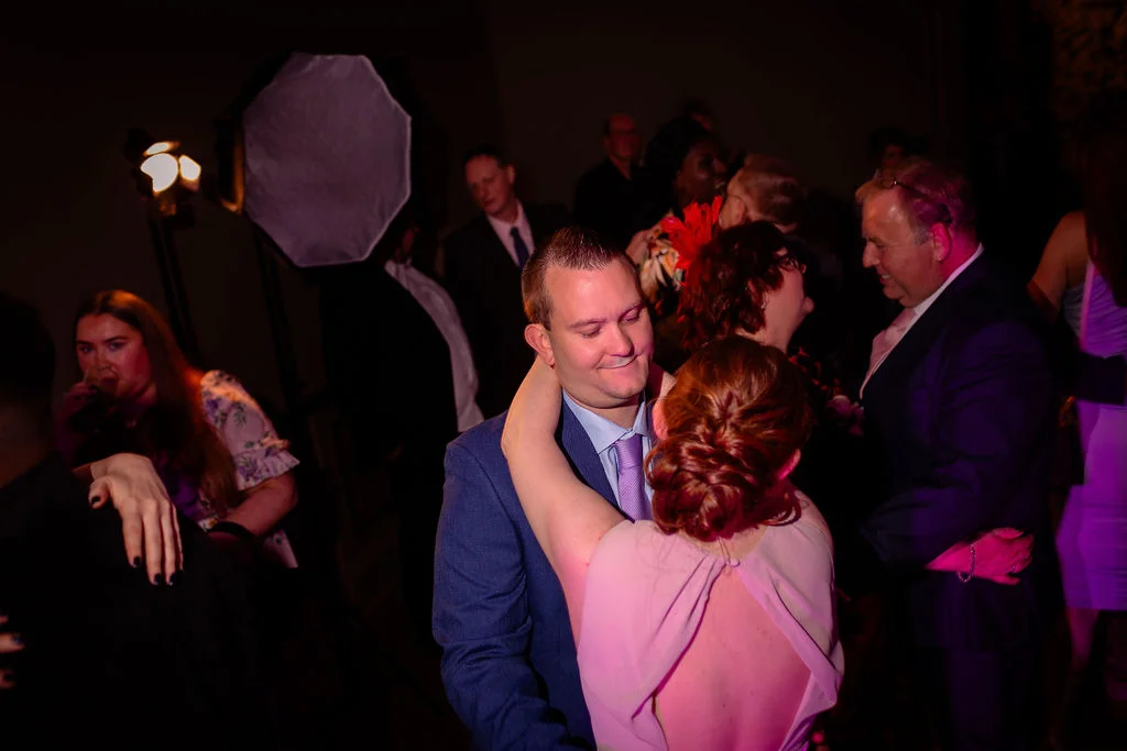 A bride and groom share a tender moment dancing together on a lively evening reception dance floor surrounded by wedding guests under ambient coloured lighting.