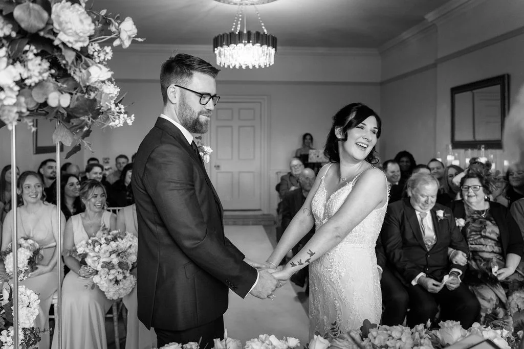A joyful bride and groom hold hands and laugh during their indoor wedding ceremony, surrounded by seated guests and elegant floral arrangements.