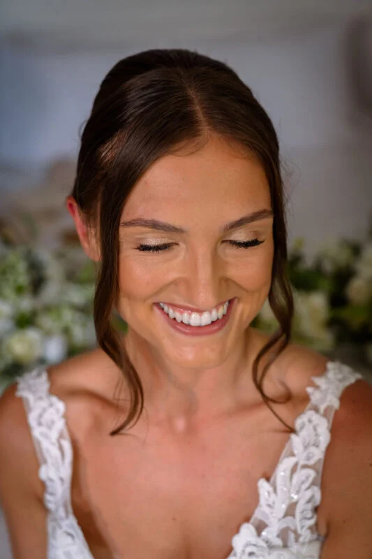 A close-up portrait of a smiling bride with her eyes closed, wearing a lace wedding dress with face-framing tendrils.
